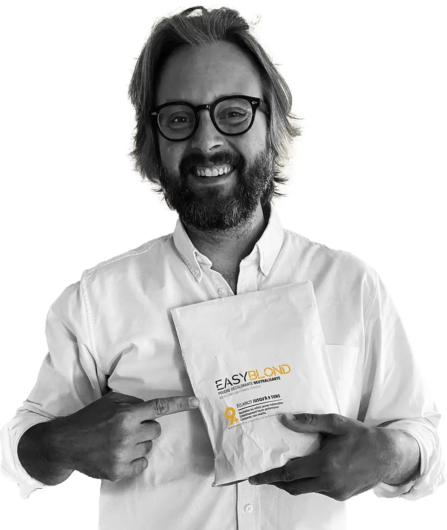 Thibaut, souriant, montre du doigt un sachet de poudre décolorante EasyBlond tenu contre sa chemise blanche