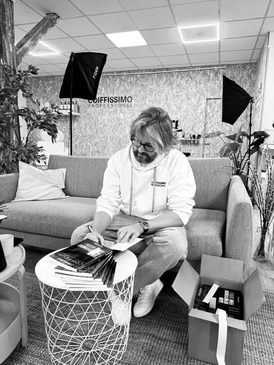 Thibaut assis sur un canapé au studio Coiffissimo, le logo Coiffissimo Professional au mur derrière lui
